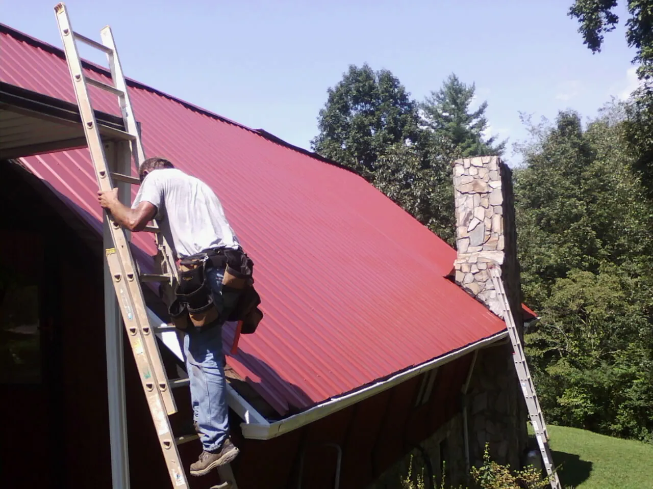Roofer on a ladder beside a red metal roof and stone chimney — Soffit & Fascia Repair by Roofing Brothers in Clayville